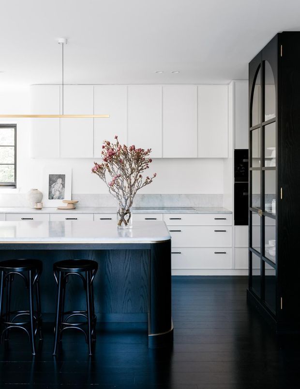 A bright contemporary kitchen with white full-height cabinetry, a pale marble-look splashback and benchtop, and a dark navy or black timber island with integrated seating. The kitchen has dark timber flooring, black stools, a slim linear pendant, and a tall black-framed glazed pantry or display door with arched detailing. A vase of flowering branches sits on the island.