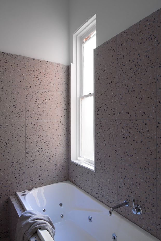 A compact bathroom with a white built-in spa bathtub beneath a narrow vertical window. The walls are clad in large square pinkish-grey terrazzo tiles with dark aggregate, paired with a plain white ceiling and white window frame. Brushed metal bath fixtures are visible at the tub edge, with a folded grey towel draped over the tub.