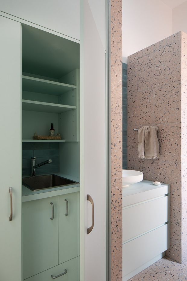 Compact contemporary bathroom with a pale green built-in utility cabinet on the left, including open shelving, a small stainless-steel sink, dark teal tiled splashback, and integrated storage. On the right is a white vanity with a vessel basin, framed by a pink terrazzo wall and matching terrazzo floor, with a neutral towel hanging on a wall-mounted rail.