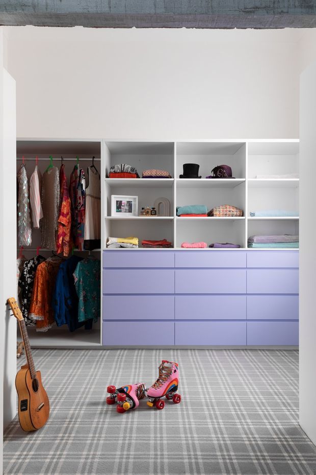 A brightly lit walk-in wardrobe or dressing room with extensive open shelving, hanging rails, and lavender built-in drawers. Colorful clothing, hats, folded garments, a guitar, and roller skates are visible, with a large grey plaid carpet covering the floor. A raw exposed concrete beam runs across the ceiling, adding an industrial architectural detail to the otherwise playful, highly organized space.