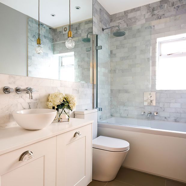 Bright, refined bathroom featuring a pale stone-look tiled shower and bath surround, glass shower screen, freestanding vessel basin on a white vanity, wall-mounted chrome fixtures, concealed-cistern toilet, and decorative pendant lights. A large mirror reflects the compact, cohesive space.