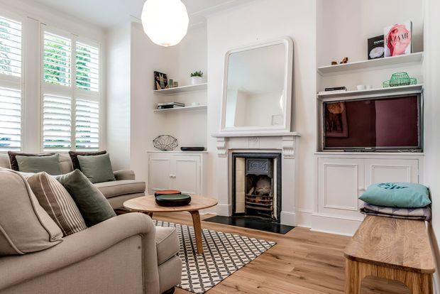 Bright, traditionally detailed living room with upholstered sofas, a timber coffee table, patterned rug, built-in shelving and cabinetry, a framed television, and a decorative fireplace with mantel and mirror. White plantation shutters and warm timber flooring create a refined, comfortable interior.