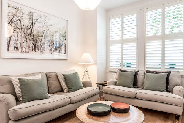Bright, welcoming living room with two upholstered sofas, layered neutral cushions, a round timber coffee table, floor lamp, wall art, and large white plantation shutters admitting abundant natural light. The palette is soft beige, cream, grey, and muted sage green, creating a refined traditional-contemporary interior.