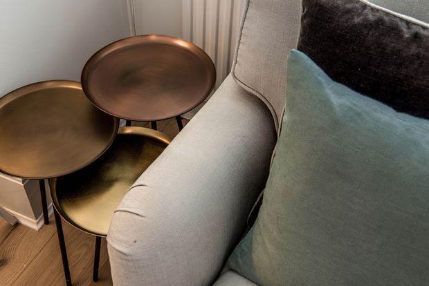 Close-up interior detail showing a light neutral upholstered armchair or sofa beside a cluster of small round nesting tables with bronze and dark metallic finishes. Teal and dark charcoal cushions add colour contrast, while a warm timber floor and softly draped curtains complete the intimate sitting area.