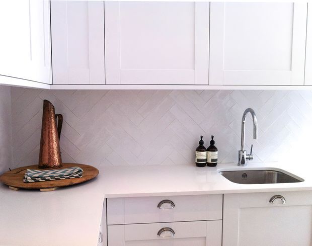 Bright, compact kitchen detail featuring white shaker-style cabinetry, a white engineered-looking benchtop, pale herringbone tile splashback, polished chrome gooseneck tapware, and a stainless-steel undermount sink. A copper decorative vessel and small accessories provide warm accents.