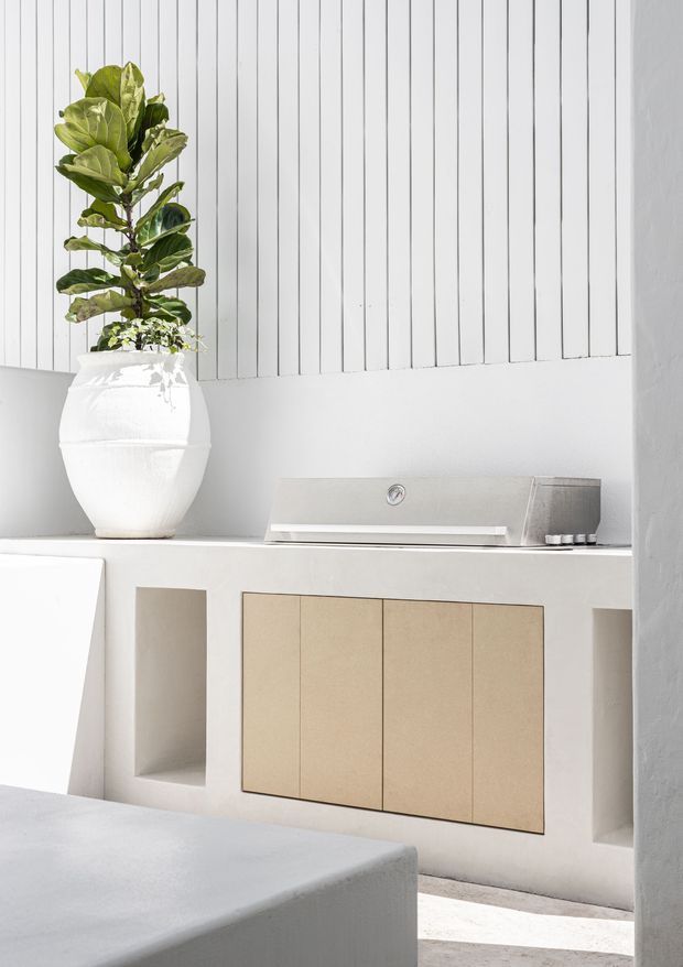 Bright minimalist interior vignette featuring a sculptural white ceramic planter with a tall leafy plant, a pale built-in sideboard with flush beige cabinet doors and open niches, and a sleek metallic linear object on top. The space uses white vertical wall panelling, pale plaster surfaces, and soft natural light.