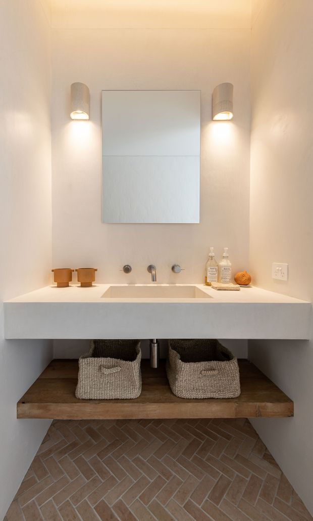 A compact, warm-toned bathroom featuring a wall-mounted white double vanity with an integrated rectangular basin, brass-toned tapware, a large frameless mirror, and paired wall sconces. Woven storage baskets sit on a substantial timber shelf below the vanity, with herringbone terracotta-toned brick flooring and softly textured light walls.