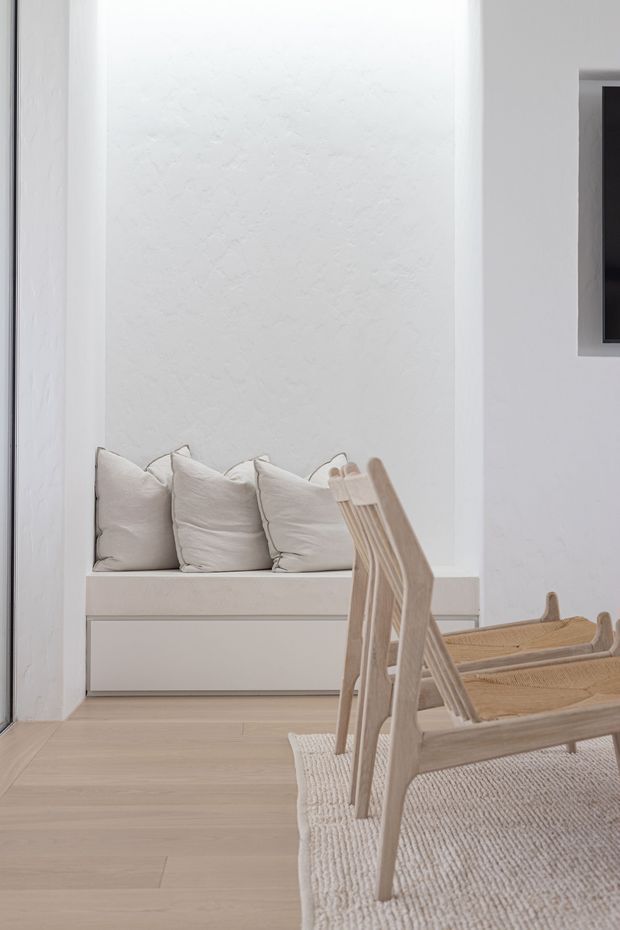 A softly lit minimalist interior corner with a built-in window seat or bench featuring white cushions, pale timber flooring, and light natural-wood sling chairs positioned on a textured neutral rug. White textured plaster walls and simple built-in joinery create a calm, pared-back aesthetic. A partial dark-framed wall opening or artwork is visible at right.