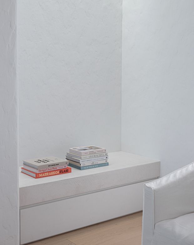 Minimalist interior vignette featuring a built-in white plastered window seat or low storage bench, lightly textured white walls, pale timber flooring, and neatly stacked books. The edge of a white upholstered armchair or sofa is visible in the foreground.