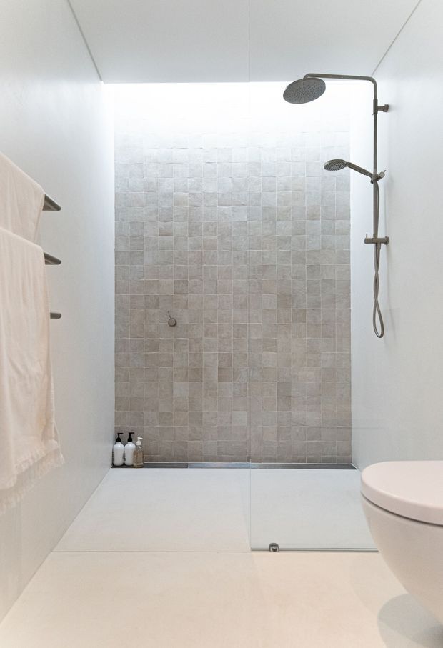 Bright minimalist bathroom with a walk-in dual-shower enclosure. The shower walls feature pale square stone-look tiles, with white walls and floor, a frameless glass screen, dark metal shower fittings, a wall-mounted towel rail, and a partially visible toilet.