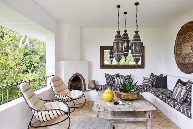 Bright, open-sided living room or sunroom with white walls, a tiled floor, built-in corner fireplace, patterned built-in seating, two cushioned black metal lounge chairs, a rustic timber coffee table, and ornate hanging lanterns. The space opens onto leafy greenery through a large balcony-like opening and features eclectic Moroccan-inspired decorative elements.