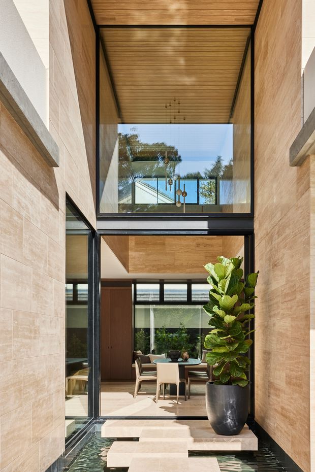 A tall, refined interior courtyard or entry passage with full-height black-framed glazing, warm timber-lined ceiling and upper wall surfaces, light stone wall cladding, and shallow water surrounding stepping-stone pavers. The view leads into a bright dining area with a round table and chairs, while a large potted plant provides greenery in the foreground. The space has strong vertical proportions, abundant natural light, and a calm indoor-outdoor connection.