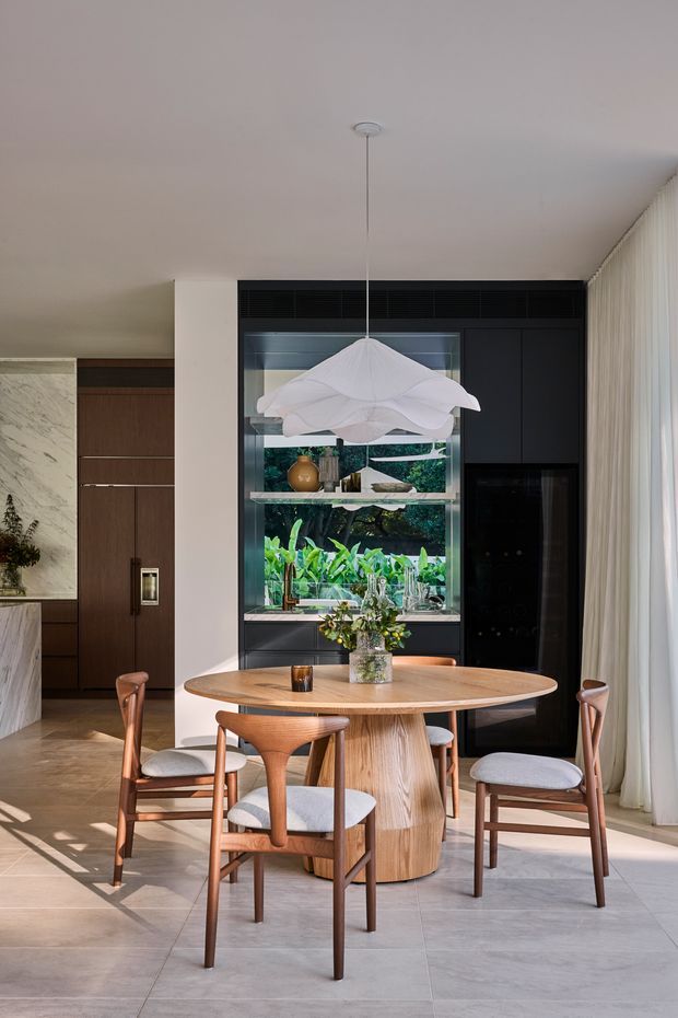 A refined dining area with a round light-wood pedestal table and four upholstered timber dining chairs. A sculptural white pendant hangs above the table. Behind it is a dark built-in wall with black cabinetry, a large glass-fronted display or garden-facing opening, and a tall dark appliance or storage unit. The room features large pale stone-look floor tiles, warm timber joinery, white walls, full-height sheer curtains, and abundant greenery visible beyond the glazing.