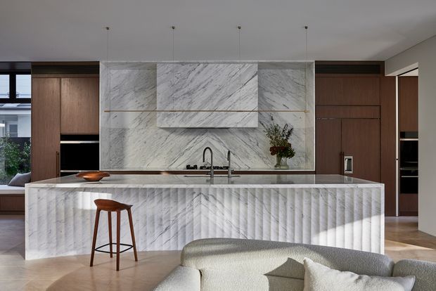 A refined contemporary kitchen with a large marble island and matching full-height marble splashback, featuring dramatic veining and a sculptural integrated rangehood surround. Warm dark timber cabinetry frames the kitchen, while slim pendant lights, brass-toned detailing, and a curved upholstered sofa create a sophisticated open-plan setting.