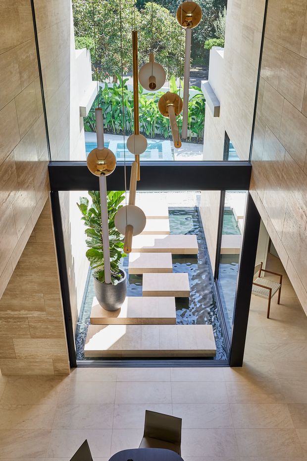 A tall, light-filled interior overlooking a narrow indoor-outdoor water feature or courtyard. Full-height black-framed glazing is set between warm timber wall panels, with floating pale stone stepping slabs over dark water, tropical planting, and sculptural spherical pendant lights suspended through the void. A small dining setting is partially visible in the foreground, and a chair sits beside the glazing.