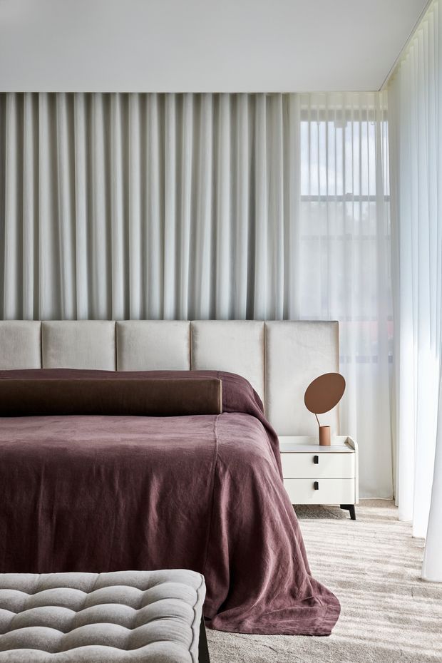 A softly lit contemporary bedroom featuring a large upholstered bed with a segmented cream headboard, layered plum and brown bedding, and a tufted upholstered bench in the foreground. Full-height sheer and heavier curtains cover tall windows, while a small white bedside table holds a simple round mirror or lamp. The room has a calm, refined, neutral-toned interior with plush textiles and carpeted flooring.