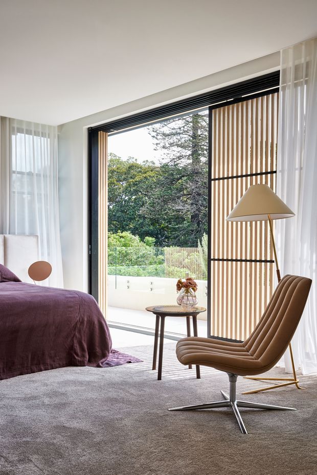 A bright, refined bedroom with a low-profile bed dressed in deep plum bedding, a grey carpet, and a sculptural brown lounge chair beside a small round side table. Full-height sheer curtains frame a large black-framed glazed opening to a terrace and garden, with a vertical timber slat privacy screen and a contemporary floor lamp. The room combines soft neutral finishes, natural timber detailing, and strong indoor–outdoor connection.