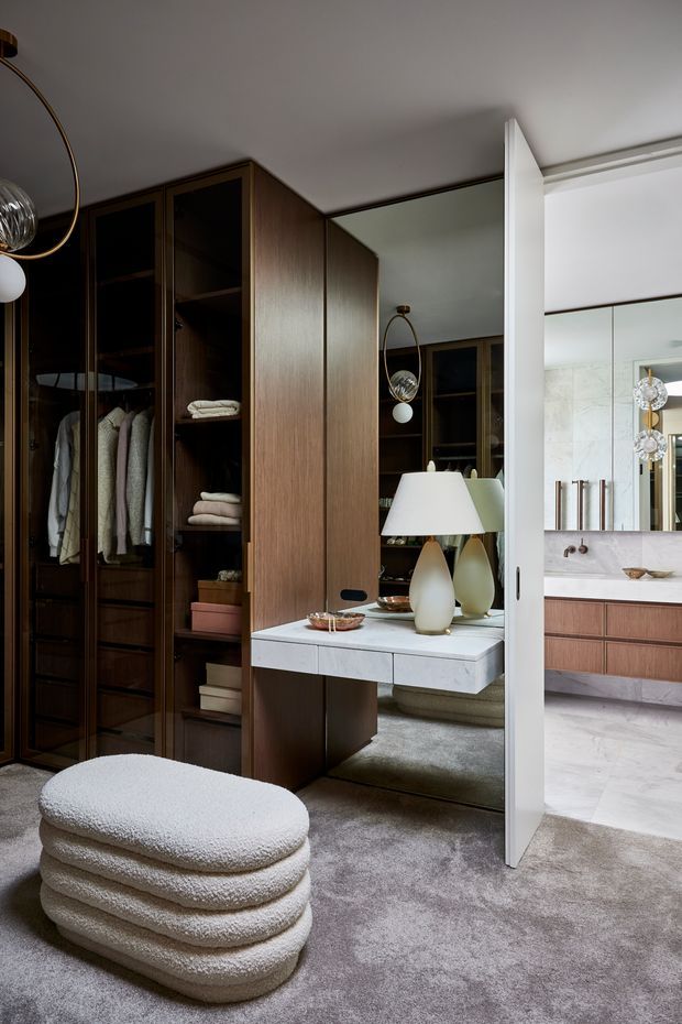 A refined walk-in wardrobe with dark timber cabinetry, glass-fronted hanging and shelving storage, plush grey carpet, and a mirrored dressing area with a floating stone-topped console. A sculptural lamp and upholstered ottoman add soft, luxurious detailing. A bathroom is visible through the open doorway.