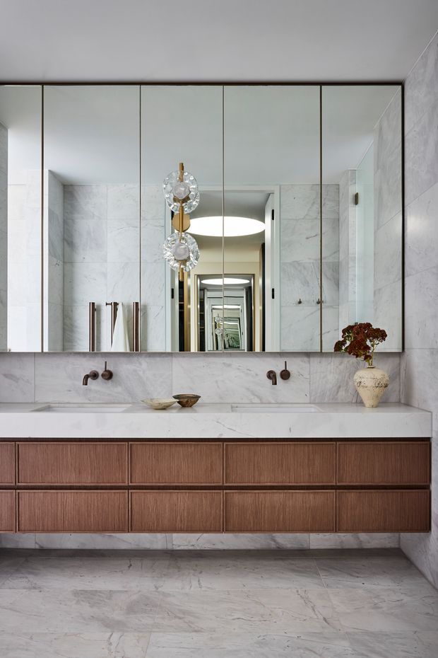 A refined contemporary bathroom featuring a long double vanity with fluted warm timber cabinetry, a pale marble benchtop and matching full-height marble wall and floor surfaces. A large wall-to-wall mirrored cabinet reflects the room and a sculptural pendant light, while brushed dark-metal wall-mounted tapware and understated accessories add contrast.