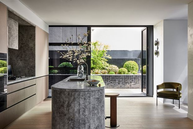 A refined contemporary kitchen with a large dark stone waterfall island, integrated cabinetry, and a sculptural tap. Full-height glazing and an open door connect the kitchen to a private courtyard with clipped shrubs and dark masonry. A sculptural lounge chair and timber stool add restrained furnishings to the bright, minimalist space.