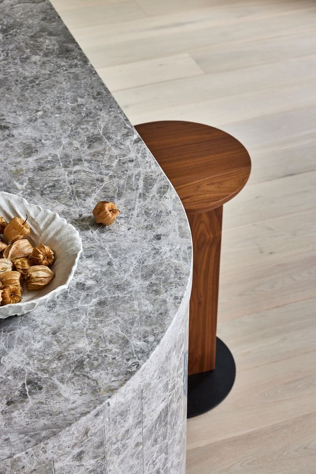 Close-up interior detail of a curved kitchen island or counter clad in highly veined grey stone, with a pale timber floor and a small round dark walnut table or stool beside it. A sculptural light ceramic bowl with decorative objects sits on the stone surface. The image highlights refined material contrast and curved joinery.