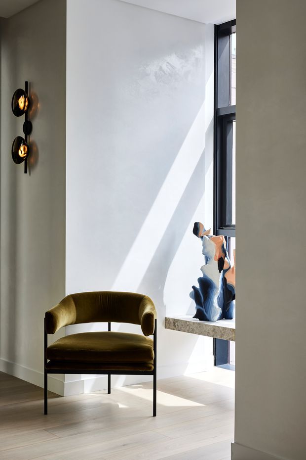 A bright, refined interior corner featuring a sculptural olive-green upholstered armchair beside a tall black-framed window. Strong sunlight casts a diagonal beam across smooth white plaster walls and pale timber flooring. A stone window ledge displays an abstract blue and cream decorative object, while a dark sculptural wall light provides a contrasting accent.