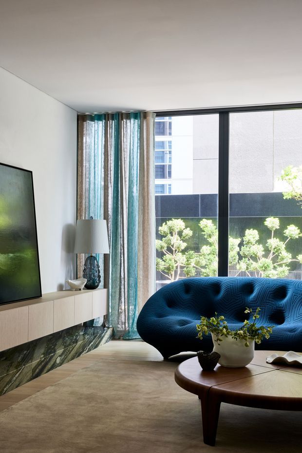 A refined contemporary living room with a deep blue sculptural sofa, round wooden coffee table, large floor-to-ceiling windows, patterned sheer curtains, a wall-mounted television, and a pale floating media console. The room has a neutral palette with blue accents, abundant natural light, and a view toward landscaped planting and neighbouring urban buildings.