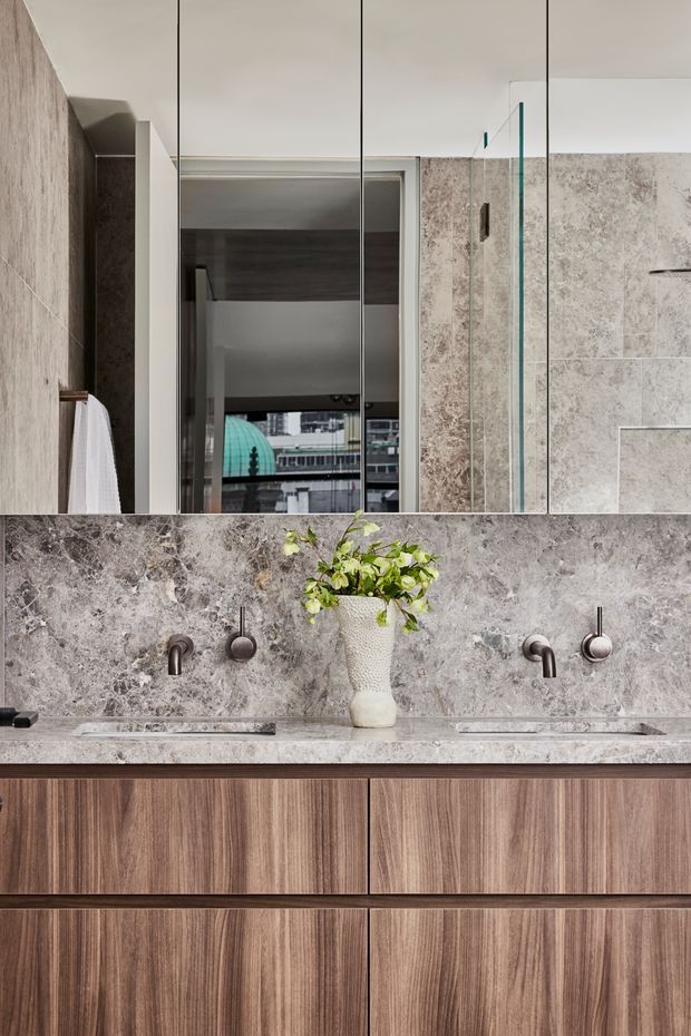 A refined contemporary bathroom vanity with a wide wall-to-wall mirror, double vessel-style wall-mounted faucets, and a long grey-veined stone benchtop and splashback. Warm timber-grain cabinetry provides contrast beneath the stone surface. A frameless glass shower enclosure is visible at right, with an urban building reflected in the mirror.