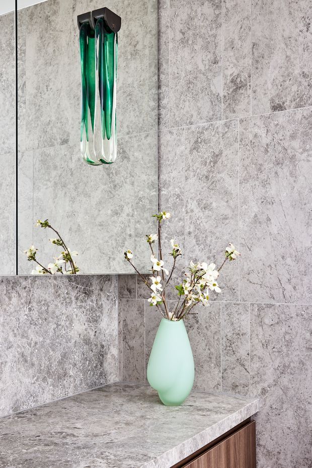 Close-up of a refined bathroom vanity or washroom feature, with grey veined stone-look wall tiles and matching benchtop, a large frameless mirror, dark timber-toned vanity cabinetry, and a sculptural mint-green vase with white branches. A suspended green glass pendant adds a vivid decorative accent.