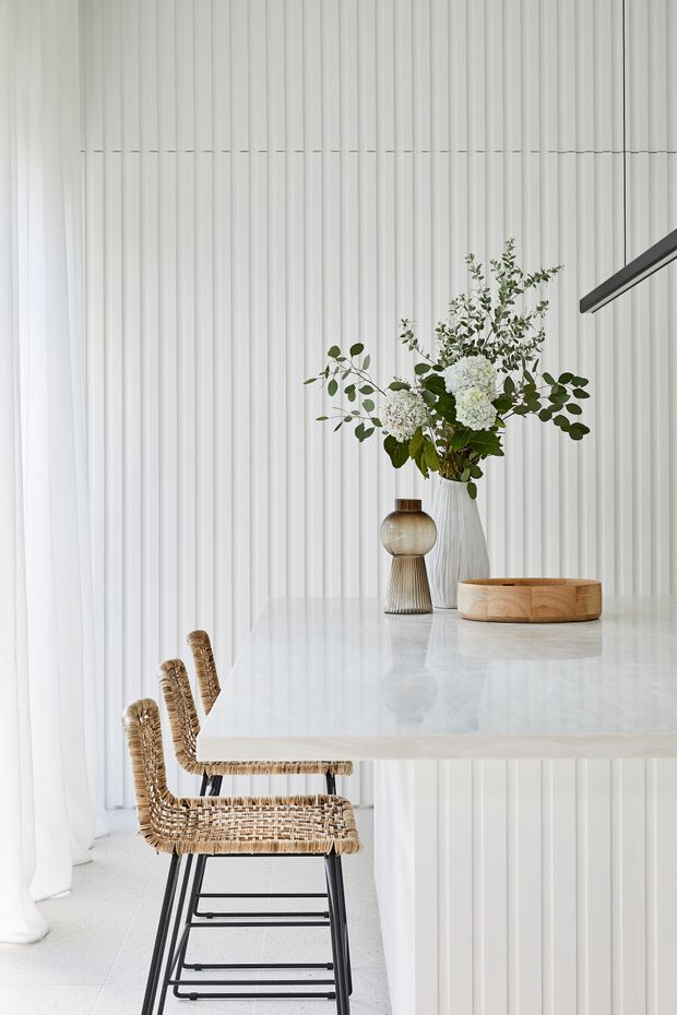 A bright, refined kitchen or dining area with a large pale stone island or countertop, white vertically ribbed wall and island paneling, and woven natural-fibre bar stools with black metal legs. A sculptural floral arrangement and decorative vessels sit on the counter, with sheer white curtains along the left side and a slim dark linear pendant light overhead.