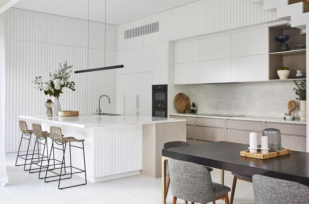 Bright open-plan kitchen and dining area with a large white island, integrated cabinetry, ribbed wall detailing, light stone benchtops, timber-toned lower cabinets, and a dark dining table with upholstered chairs. A staircase and curated shelving are visible in the background. The space has a refined, contemporary minimalist aesthetic with a light neutral palette.