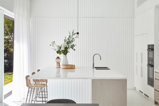 Bright minimalist kitchen with a large white island, integrated sink and tall curved tap, pale timber cabinetry, and a vertically ribbed white feature wall. A woven bar stool, simple ceramic vessels, greenery, and sheer curtains add restrained natural accents. Full-height glazing opens to a garden exterior.