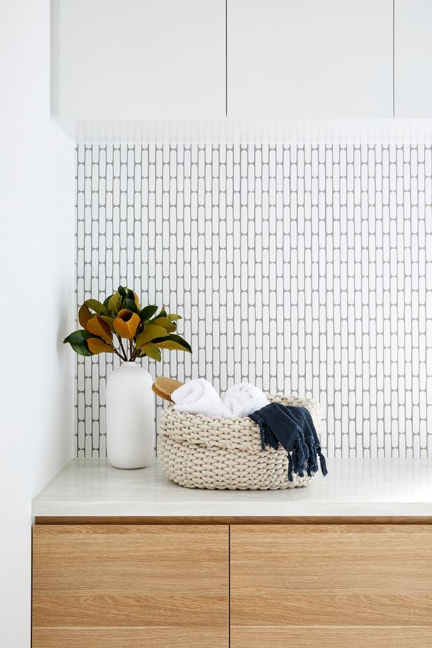 A bright, minimalist built-in vanity or storage console with pale timber drawers, a white benchtop, and a white patterned tile backsplash. A woven basket containing folded towels and a dark blue textile sits beside a white vase with leafy branches, adding natural texture and colour.