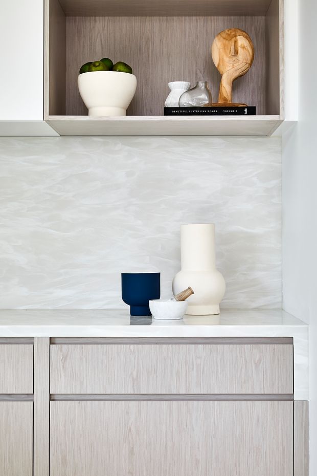 Close-up of a minimalist built-in kitchen or living-room display niche with pale wood-grain cabinetry, white shelving, and a softly textured white backsplash or wall finish. The open upper shelf contains a white bowl with green fruit, a wooden serving spoon, small white vessels, and a book. Below, a white counter displays sculptural ceramic vases and a dark blue cup, with pale wood drawers and cabinetry beneath.