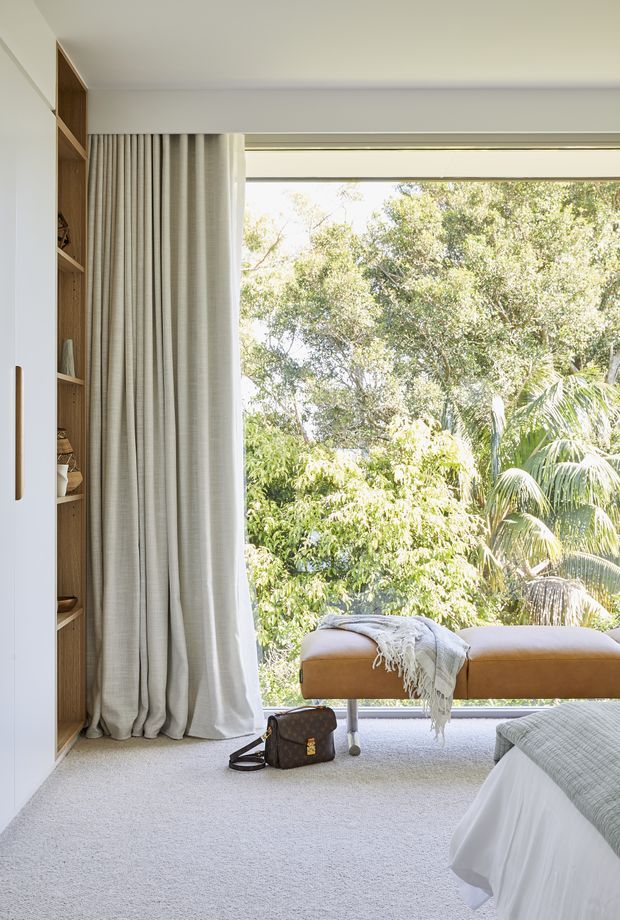 Bright, airy bedroom with a large floor-to-ceiling window overlooking lush greenery. The room features soft neutral carpeting, full-height beige curtains, white built-in cabinetry with open timber shelving, and a tan upholstered bench at the window. Part of a neatly made bed is visible in the foreground, creating a calm, refined contemporary interior.
