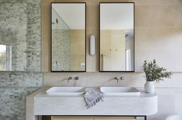 Contemporary bathroom featuring a double vanity with two countertop basins, wall-mounted taps, frameless rectangular mirrors, a pale stone-look vanity top and splashback, and a textured patterned glass shower enclosure. The space uses soft neutral tones with black mirror frames and minimal styling.