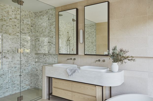 Contemporary bathroom featuring a double vanity with pale timber drawers, a white integrated benchtop, wall-mounted basin fixtures, framed mirrors, and a frameless glass shower enclosure. The shower walls use a mottled light stone or mosaic finish, while the main walls are clad in large warm beige tiles. A freestanding bathtub is partially visible in the foreground.