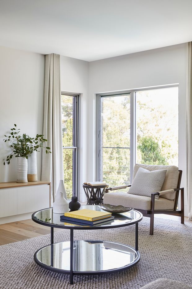 Bright, refined living room with a large round two-tier mirrored coffee table, upholstered lounge chair, textured neutral rug, pale timber flooring, pleated full-height curtains, and expansive windows overlooking greenery. A ribbed built-in cabinet and understated decorative objects add texture while maintaining a calm contemporary feel.