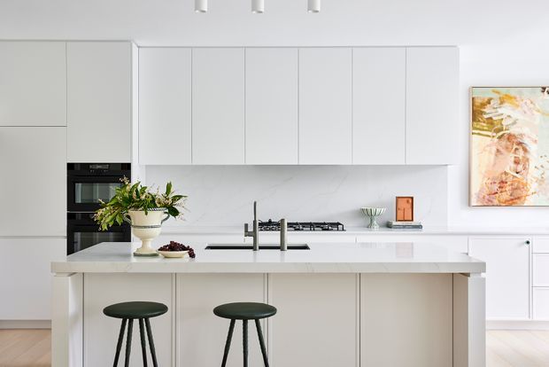 Bright, contemporary white kitchen with a large central island, integrated cabinetry, built-in oven, gas cooktop, undermount sink and two dark green bar stools. The space features pale timber flooring, white cabinetry, a light stone-look benchtop and splashback, minimalist pendant-free ceiling spotlights, and a colourful abstract artwork.