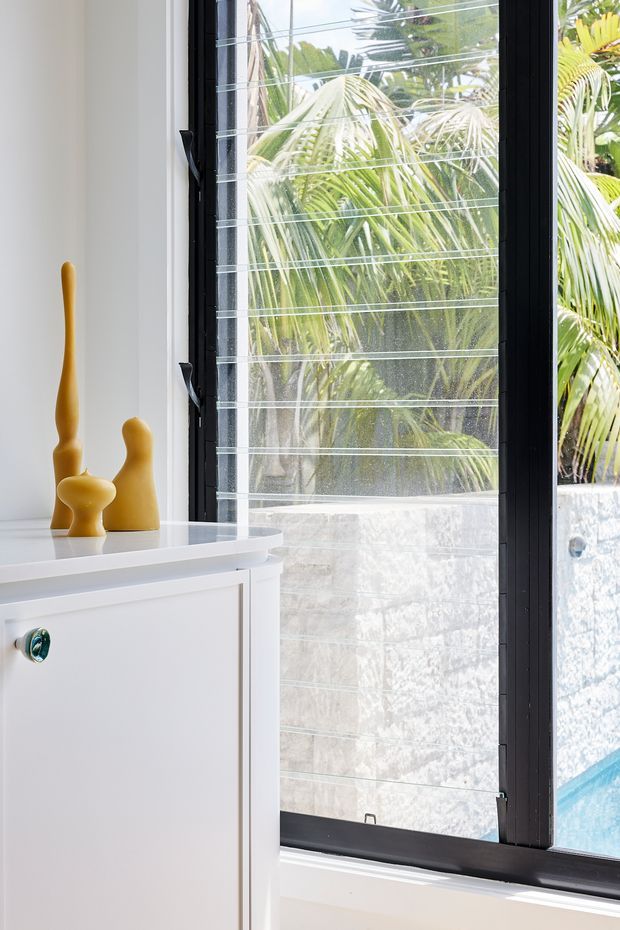 Bright interior detail featuring a white built-in cabinet with a polished white benchtop and small yellow sculptural ornaments. A large black-framed louvered window provides views of tropical palm foliage, a pale masonry wall, and a glimpse of a swimming pool outside.