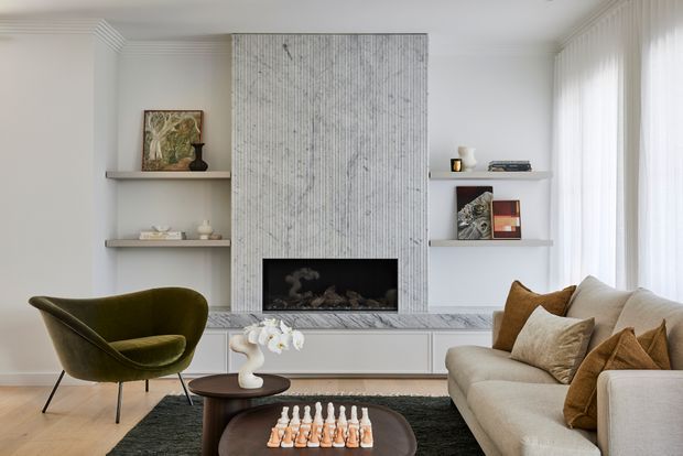 Refined contemporary living room with a sculptural marble fireplace surround and integrated firebox as the central feature. Built-in white cabinetry and open display shelving flank the fireplace, while a neutral upholstered sofa, olive green accent chair, dark timber coffee table, rug, and soft curtains create a calm, elegant setting. A chessboard and decorative objects add lived-in detail.