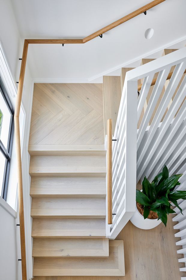 Bright contemporary residential staircase viewed from above, featuring pale timber stairs and flooring, white painted balustrades with vertical slats, and warm timber handrails. A large window provides natural light, while a potted leafy plant adds greenery beside the stairwell.