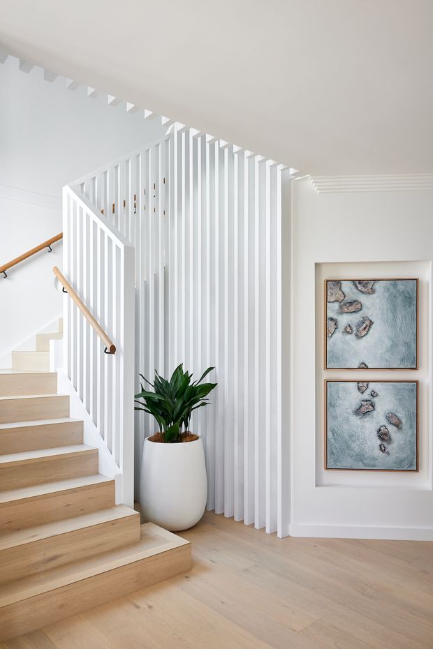Bright, refined interior view of a stairway and landing. A pale timber staircase with white balustrades and warm wood handrails rises beside a full-height vertical white slatted screen. The space has light timber flooring, white walls and ceiling, recessed artwork, and a large potted plant. Layered ceiling mouldings add a traditional architectural detail to the otherwise contemporary, minimalist setting.