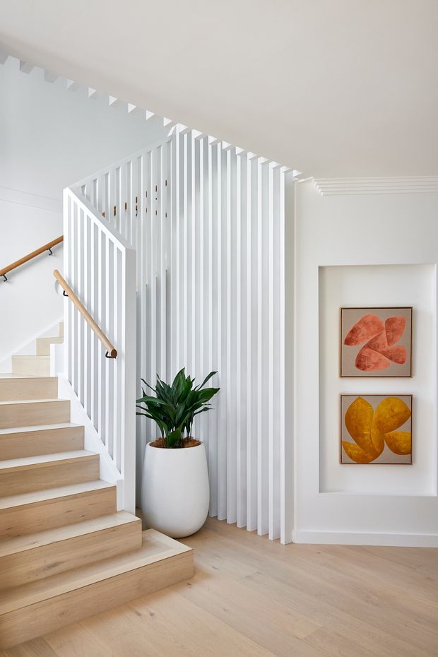 Bright, refined stairway and landing with pale timber treads and flooring, white balustrades and vertical slatted screening, warm timber handrails, and layered ceiling cornice detailing. A large potted plant and two colourful artworks sit beside a recessed wall niche.