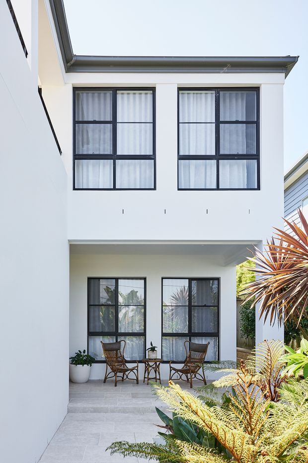 A bright, narrow residential courtyard framed by a two-storey white rendered building. The facade has large black-framed grid windows with sheer curtains, a covered ground-level patio with two woven chairs and a small table, pale stone paving, and lush tropical planting. The composition highlights the modern courtyard-facing exterior and outdoor seating area.