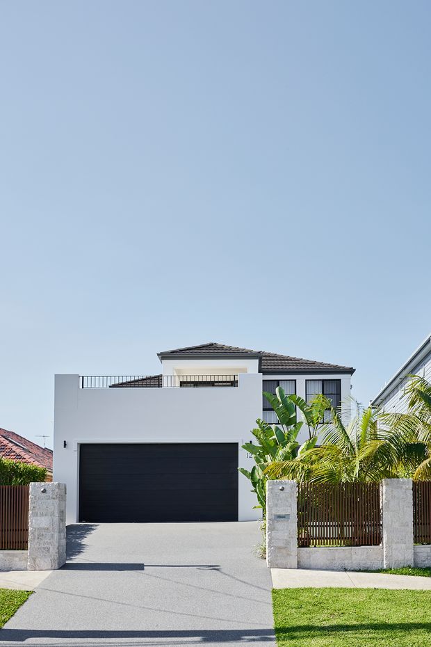 Street-front exterior view of a contemporary two-storey white-rendered residence with a dark tiled hip roof, large black garage door, upper-level balcony, black-framed windows, concrete driveway, and a low front fence with tropical planting.