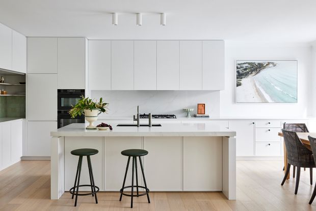 Bright, minimalist contemporary kitchen with a large white island, integrated cabinetry, built-in appliances, and a pale timber floor. Two dark stools sit at the island, while a dining area and coastal artwork are visible beyond.