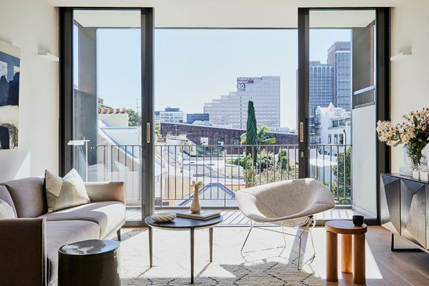 Bright, refined living room with floor-to-ceiling black-framed glazing opening to a balcony and an urban city view. The room features a neutral sofa, sculptural lounge chair, round coffee tables, a patterned rug, and a dark built-in or freestanding media console with decorative objects. Natural daylight creates an airy contemporary atmosphere.