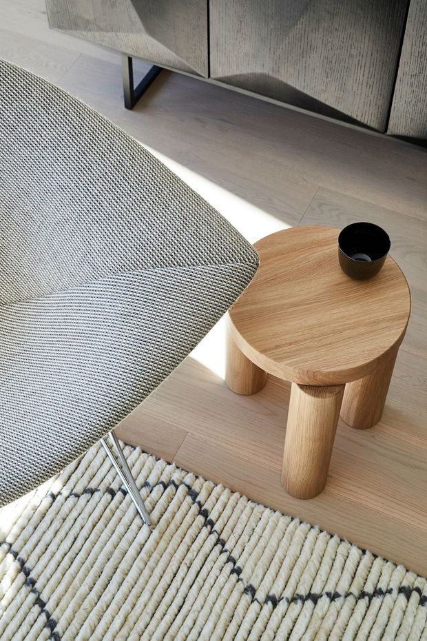 Close-up interior vignette showing a contemporary lounge chair with textured grey upholstery, a round light-oak side table with cylindrical legs, a dark ceramic vessel, pale timber flooring, a textured cream rug, and a dark timber sideboard in the background. Strong natural sunlight creates a bright, calm composition.