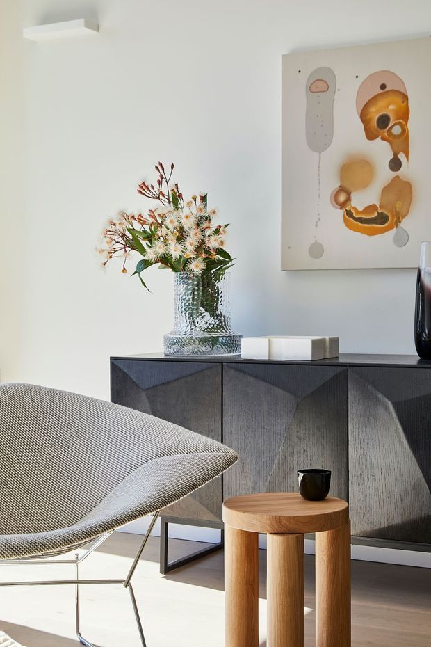 A bright, refined contemporary interior vignette featuring a sculptural upholstered lounge chair, a dark geometric-front sideboard, a round light-wood stool or side table, and a textured glass vase with native-style flowers. A large abstract artwork hangs on the pale wall above the cabinetry, with strong natural light creating crisp shadows across the light floor.